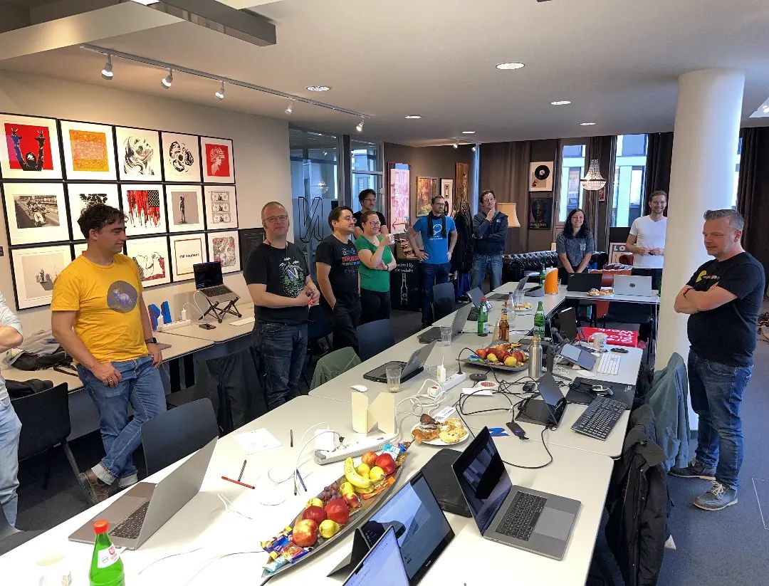 11 people from the Neos Sprint stand happily around a table, that is full of laptops, fruits and snacks.